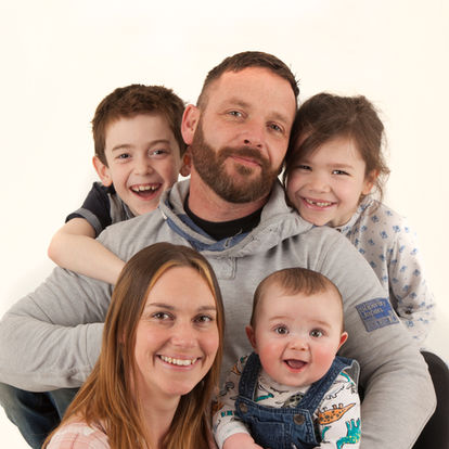 A man and woman pose closely with three smiling children, including a baby, against a plain white background. Andy Boyle Photography
