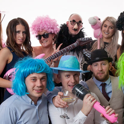 A group of people pose for a photo wearing colorful wigs, hats, and costumes, with several holding props like inflatable guitars, a microphone, and a stuffed animal. Andy Boyle Photography