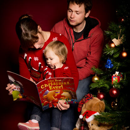 A woman reads a Christmas book to a young child while a man sits beside them near a decorated Christmas tree and stuffed toys. Andy Boyle Photography