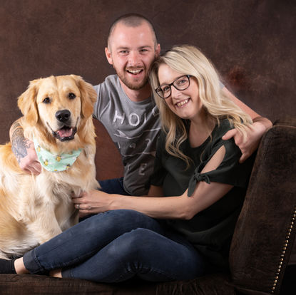 A man and woman sit on a brown couch, smiling at the camera with a large golden retriever wearing a bandana seated beside them. Andy Boyle Photography