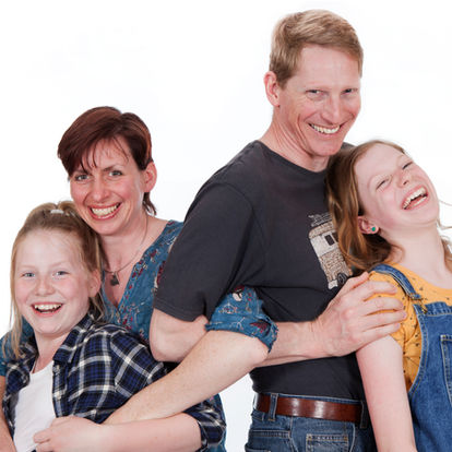 A smiling family of four poses together against a white background, with two adults and two children standing closely and laughing. Andy Boyle Photography
