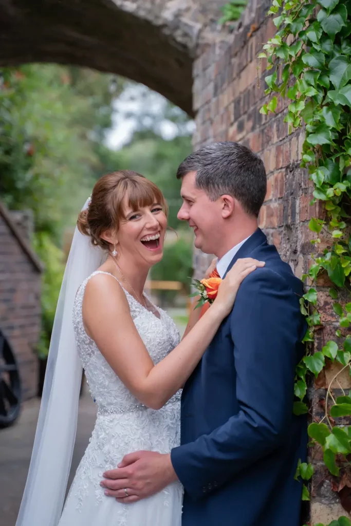 A bride in a white dress and a groom in a blue suit stand close together, smiling and laughing, next to a brick wall covered in green ivy. Andy Boyle Photography