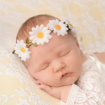 A sleeping baby wearing a white lace outfit and a white daisy flower headband rests on a lace pillow. Andy Boyle Photography