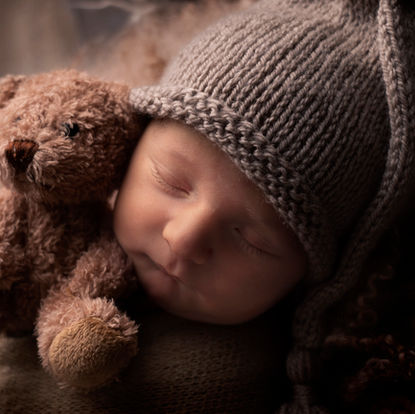 A sleeping baby wearing a knitted hat rests its head next to a soft brown teddy bear. Andy Boyle Photography