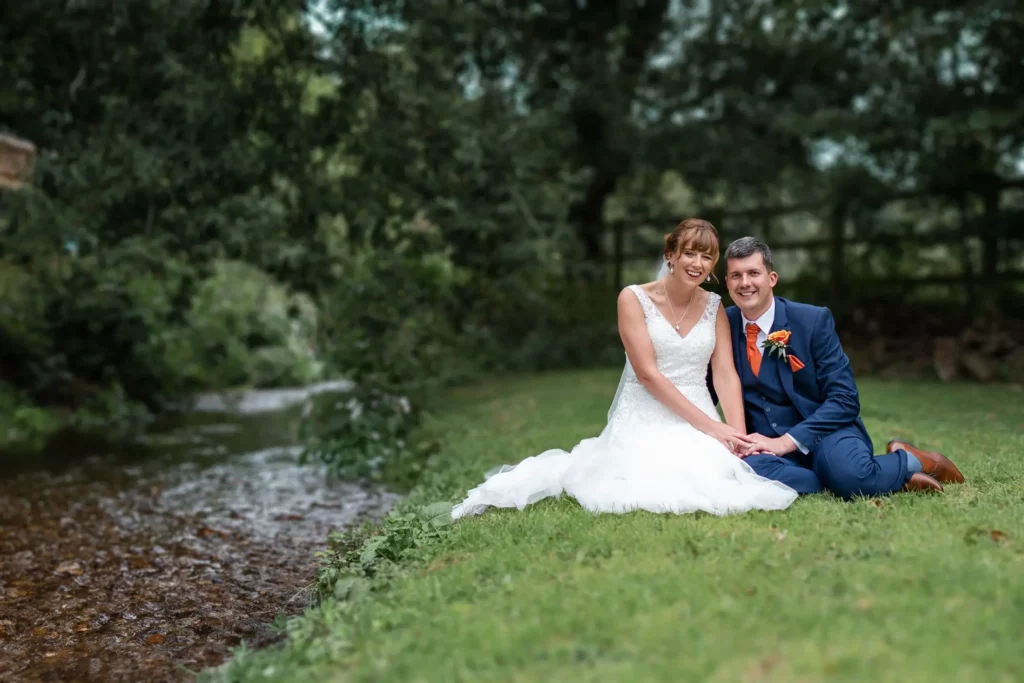 A bride and groom sit together on grass beside a stream, both smiling at the camera, with trees and greenery in the background. Andy Boyle Photography
