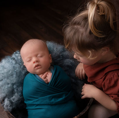A baby swaddled in a blue blanket sleeps in a basket while a young child with a ponytail looks on. Andy Boyle Photography
