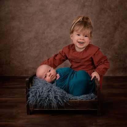A young child smiles while sitting beside a baby wrapped in a teal blanket, lying in a small wooden bed with a gray furry blanket, against a brown studio backdrop. Andy Boyle Photography