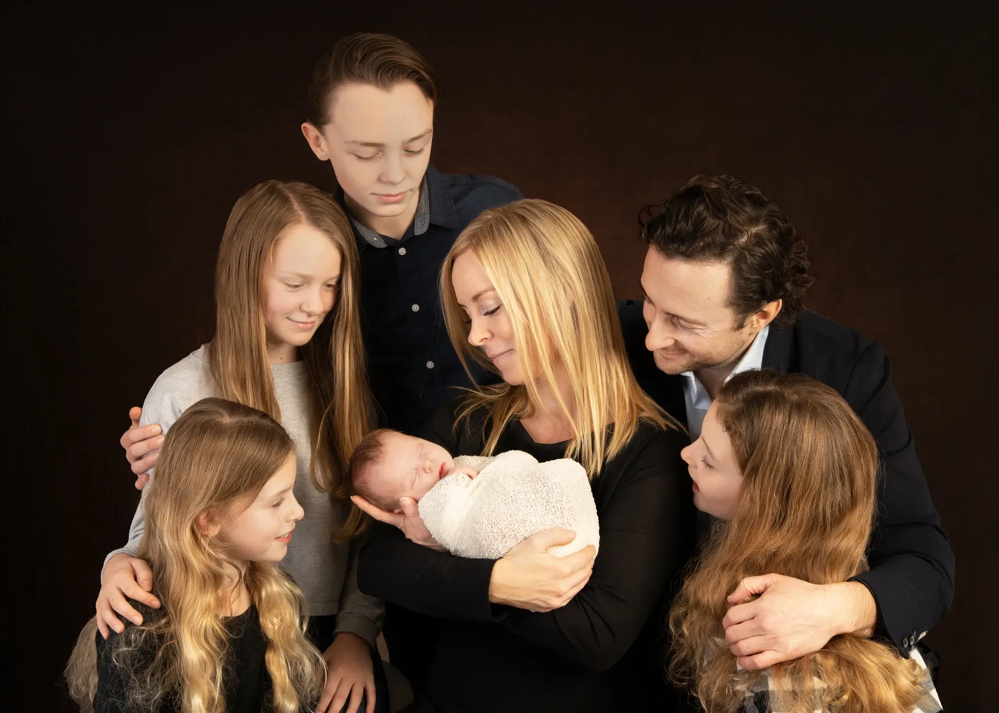 A family of seven poses together, with the mother holding a swaddled newborn while the father and four older children gather around, all looking at the baby. Andy Boyle Photography