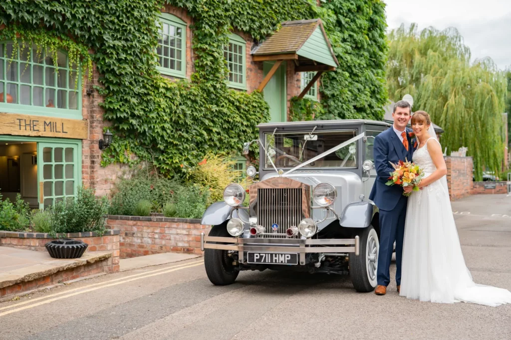 A bride and groom stand beside a vintage car decorated with ribbon, outside a building covered in ivy with a sign that reads "The Mill. Andy Boyle Photography