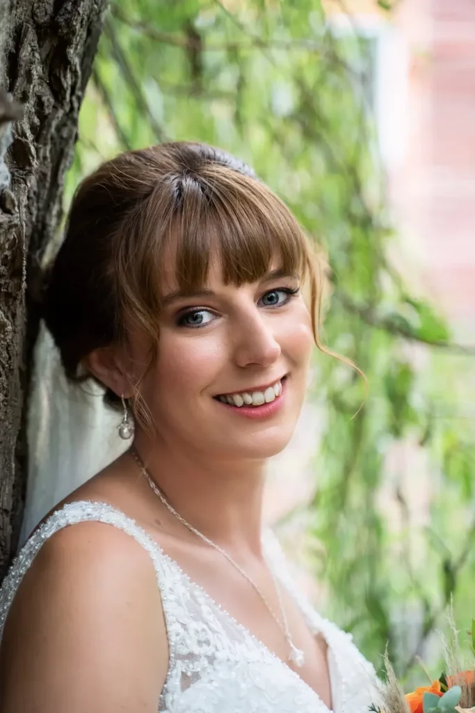 A woman in a white lace wedding dress with a veil and necklace smiles while standing outdoors next to a tree. Andy Boyle Photography