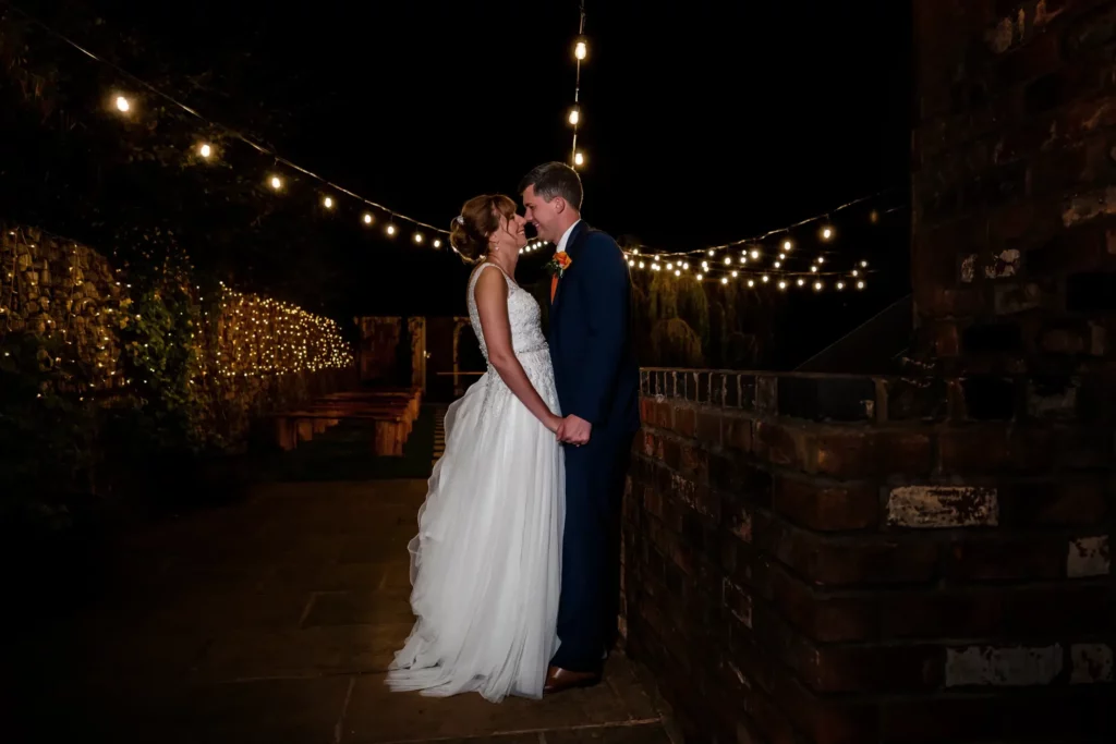 A bride and groom stand close together at night, holding hands under string lights on a brick patio, gazing at each other. Andy Boyle Photography