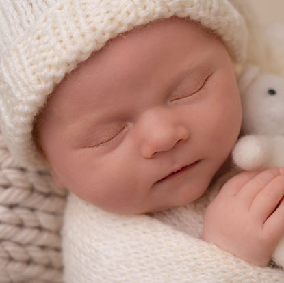 A newborn baby sleeping peacefully, wrapped in a cream-colored knitted blanket and wearing a matching hat, holding a small stuffed toy. Andy Boyle Photography