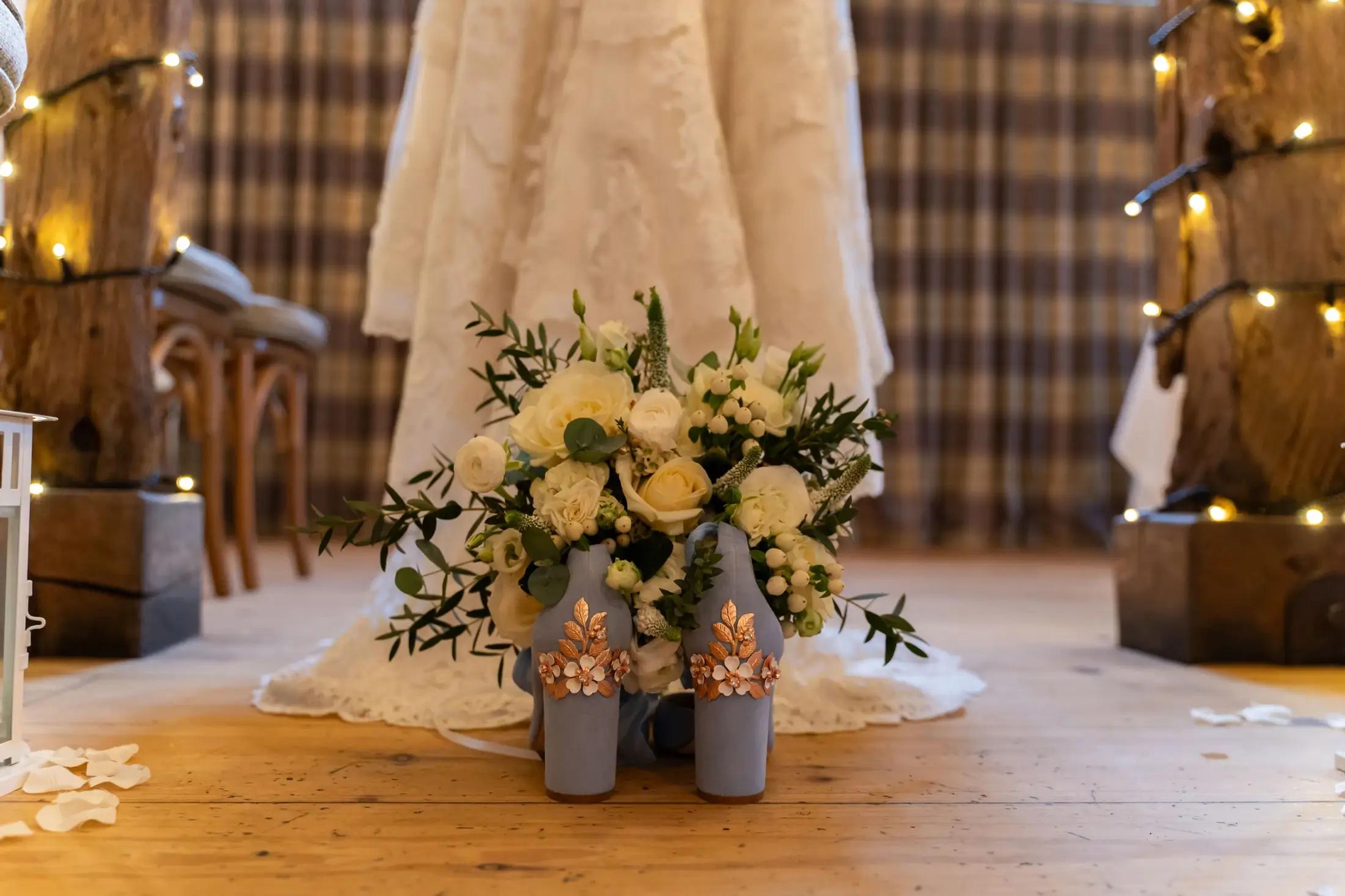 A white bridal gown with a lace hem is shown behind a bouquet of white and yellow flowers and two decorated blue bottles on a wooden floor. Andy Boyle Photography
