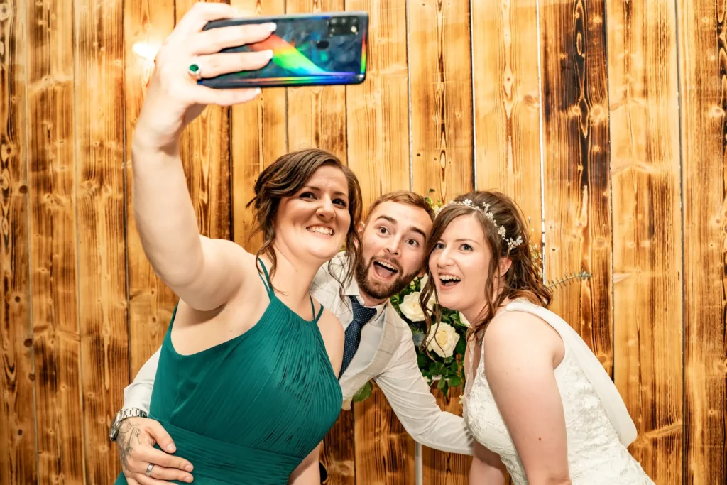 Three people, including a bride in a white dress, pose together for a selfie in front of a wooden backdrop at what appears to be a wedding. Andy Boyle Photography