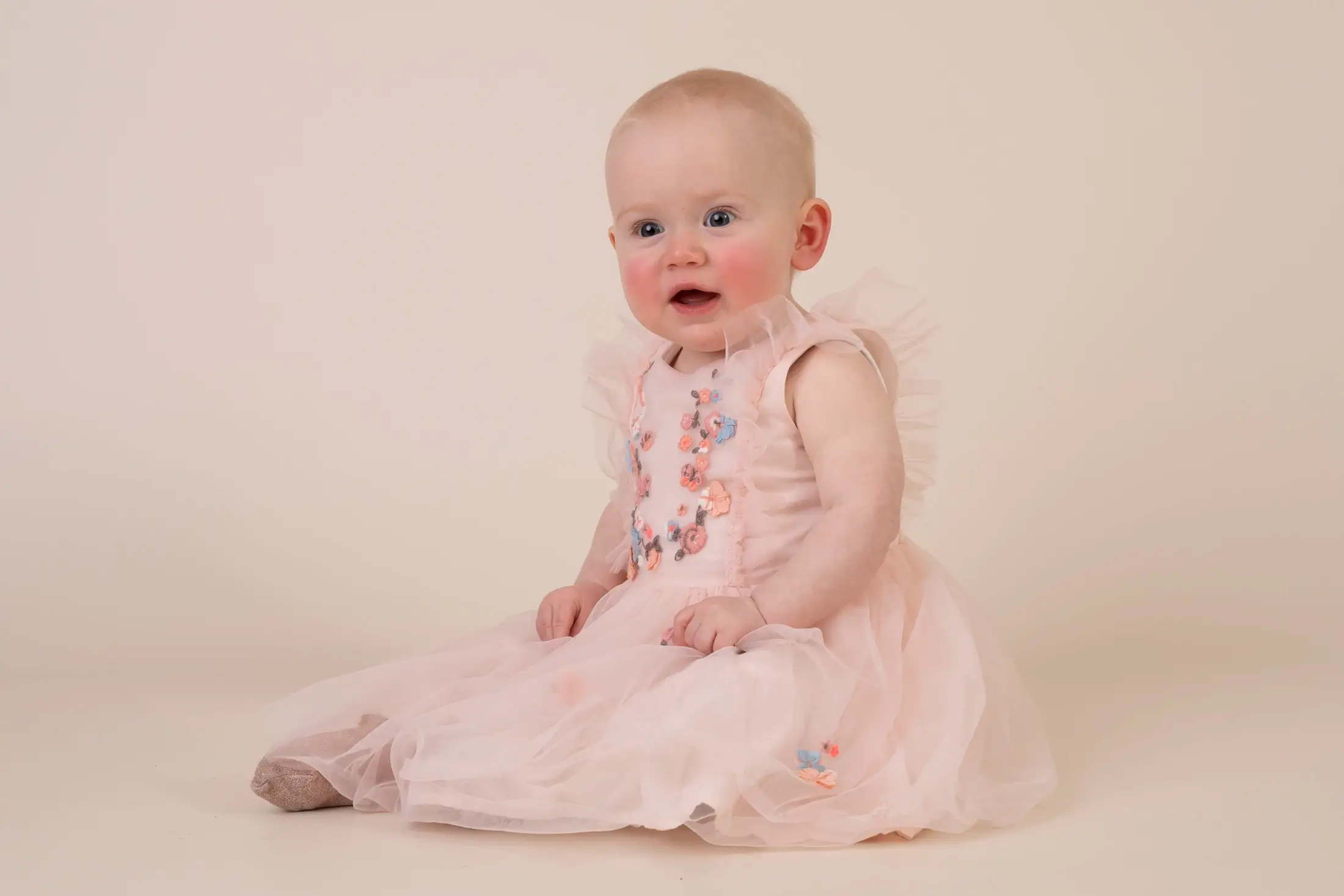 A baby wearing a light pink, frilly dress with floral embroidery sits on a neutral background, looking slightly to the side. Andy Boyle Photography