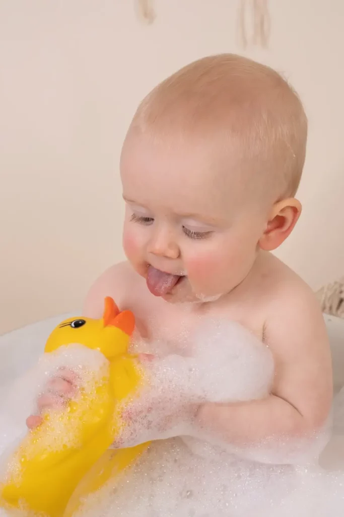 A baby sits in a bubble bath, sticking out their tongue while holding a yellow rubber duck. Andy Boyle Photography