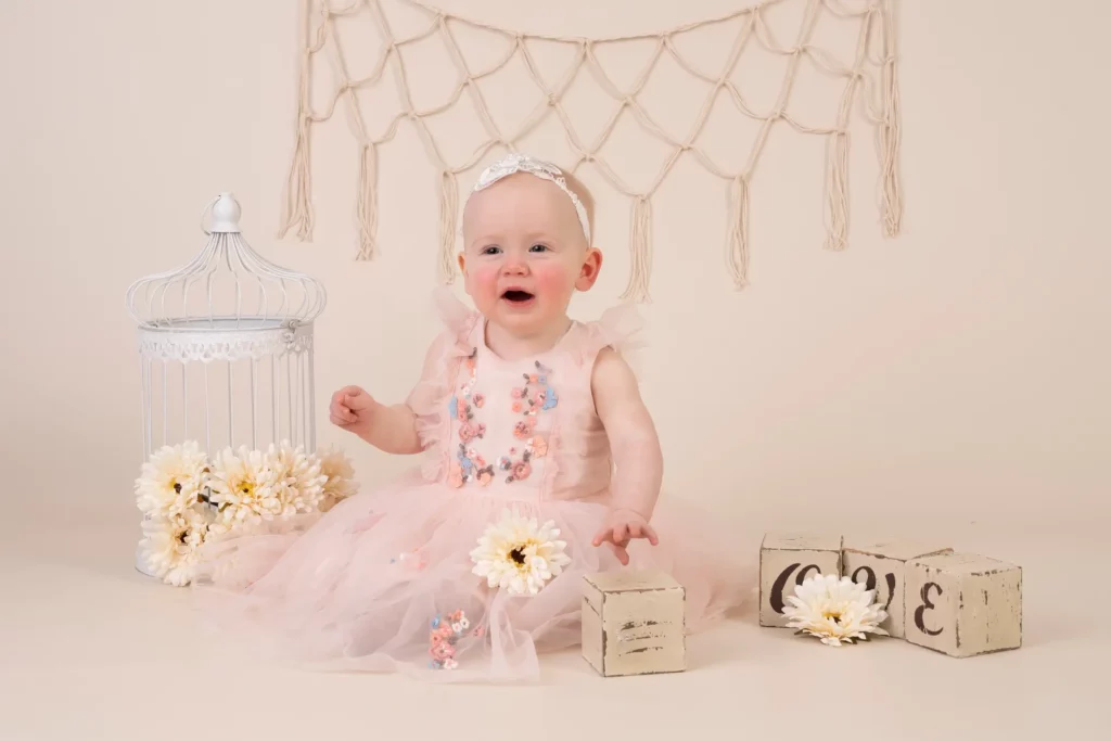 A baby in a pink dress sits on the floor with flowers, wooden blocks, and a white decorative birdcage, against a beige background with a macramé wall hanging. Andy Boyle Photography