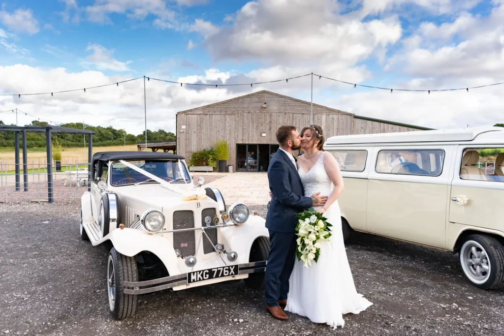 A bride and groom kiss beside a vintage white car and a cream van in front of a rustic wooden barn under a partly cloudy sky. Andy Boyle Photography